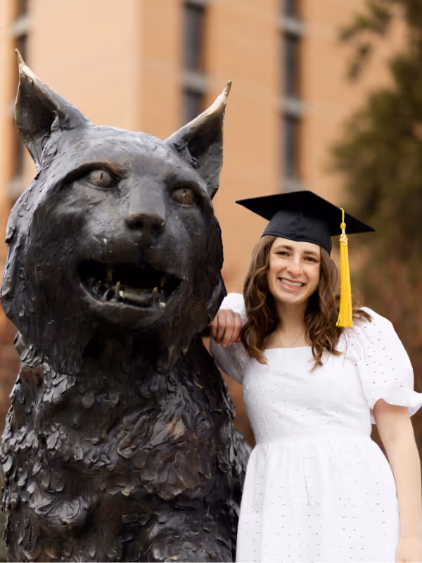 Graduate in cap and gown posing with Spirit the Bobcat statue at Montana State University during graduation