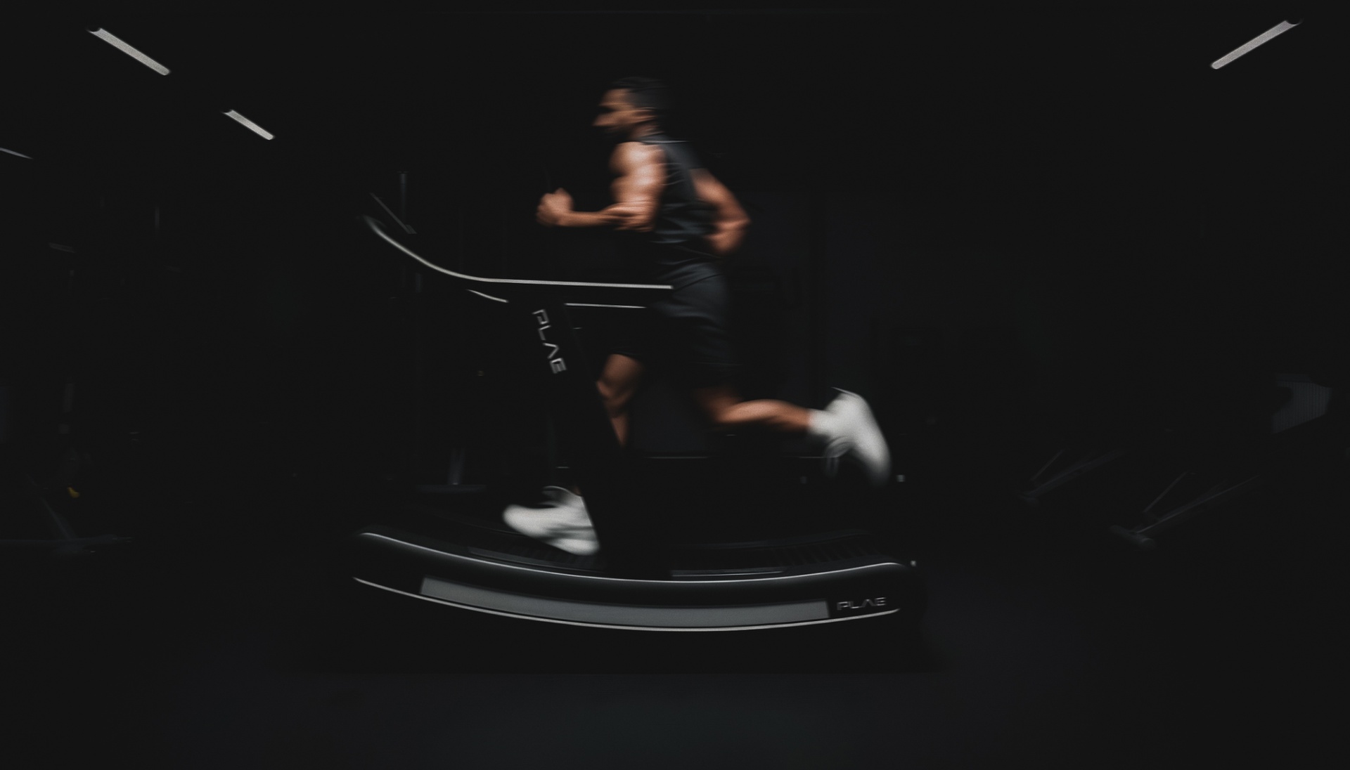 Blurred motion of a person running on a PLAE Arc curved treadmill in a dimly lit gym.