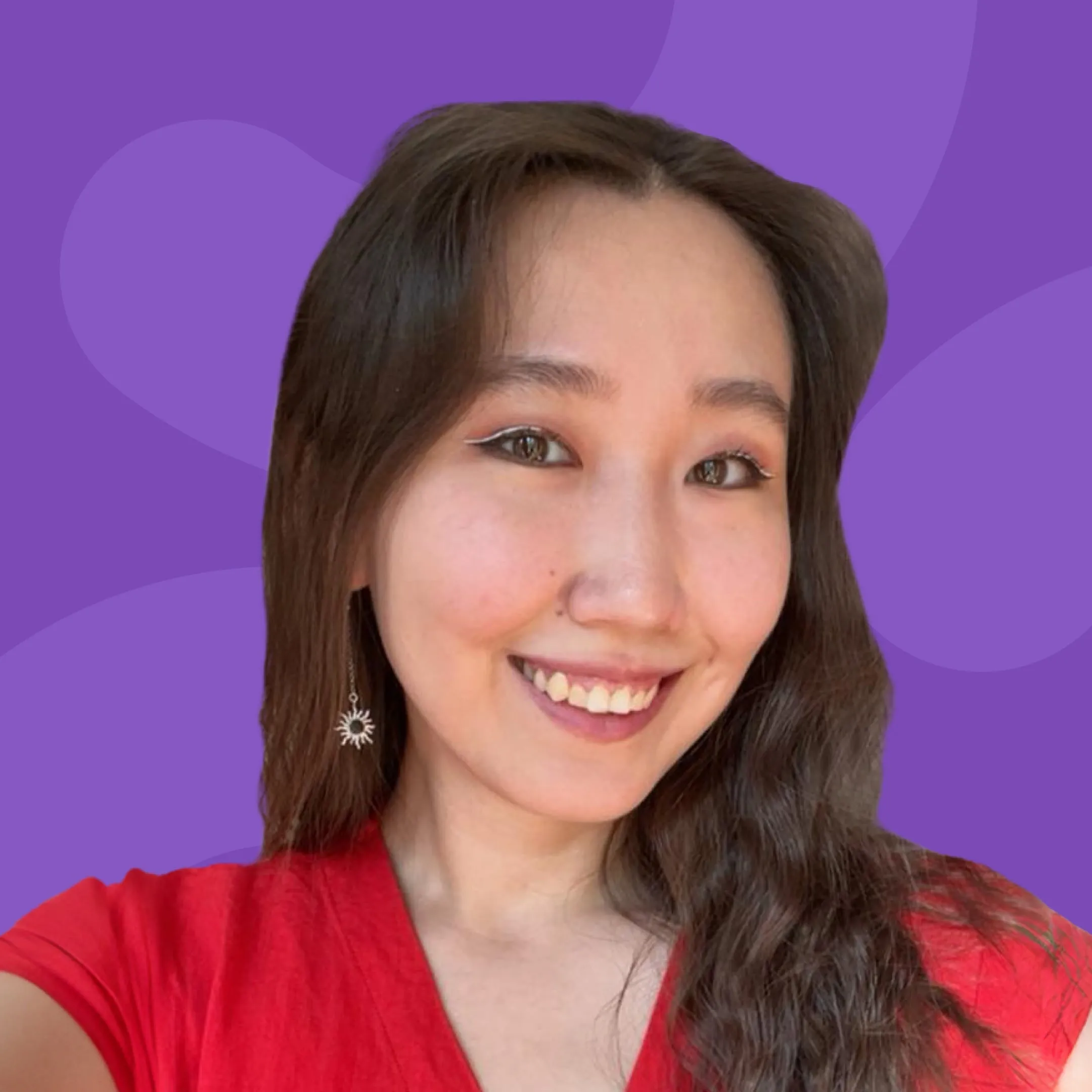 Smiling woman with long dark hair wearing a red top and sun-shaped dangling earrings against a purple background.