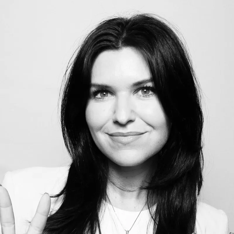 Black and white portrait of a woman with long dark hair smiling and making a peace sign with her hand.