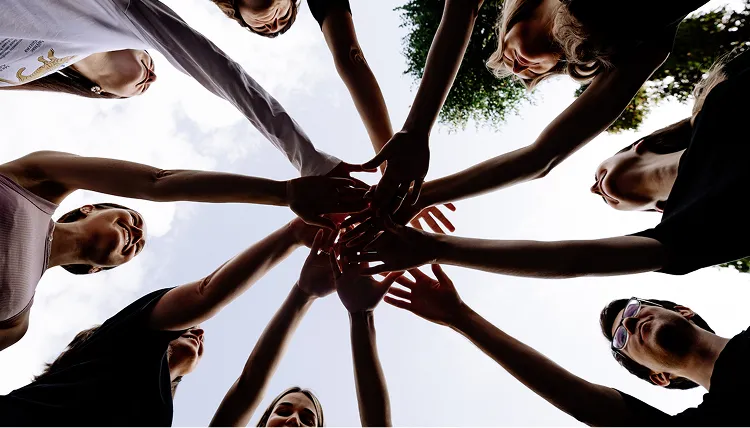 Group of people standing in a circle with hands joined together in the center against a cloudy sky.
