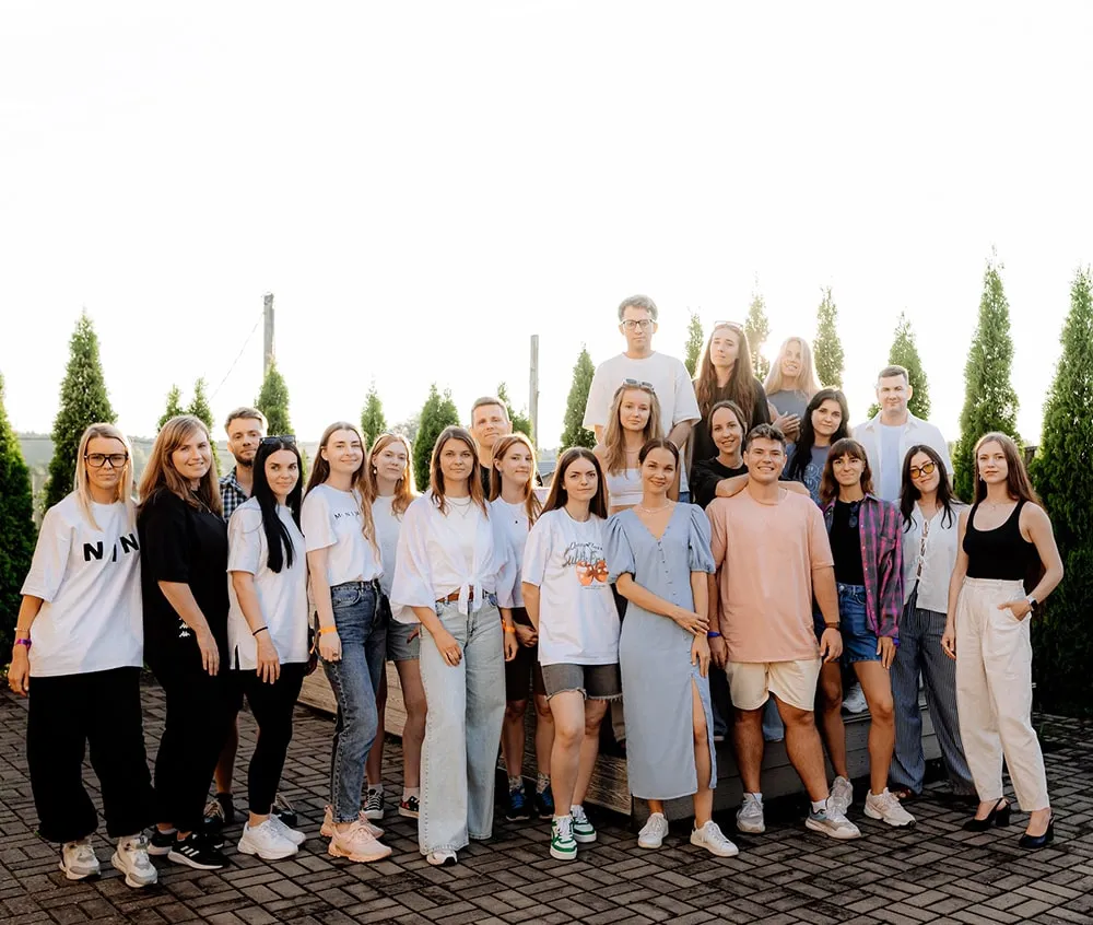 Group of 23 young adults posing outdoors on a paved area with tall green trees in the background.