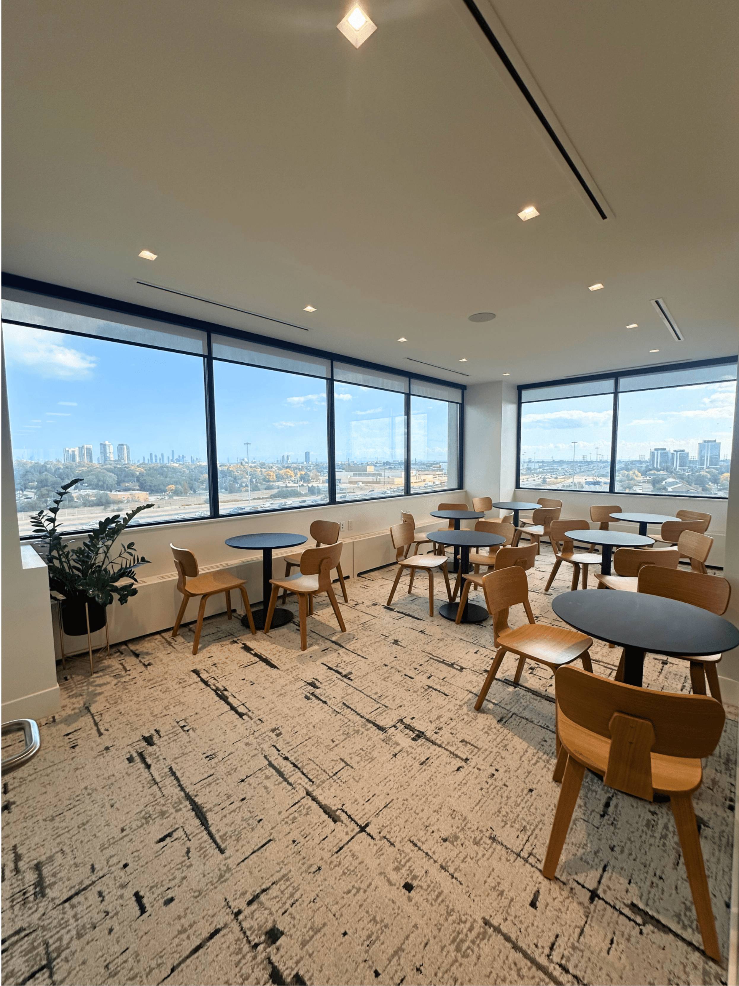 Modern event space with round black tables, wooden chairs, large windows offering a cityscape view, and patterned carpet flooring.