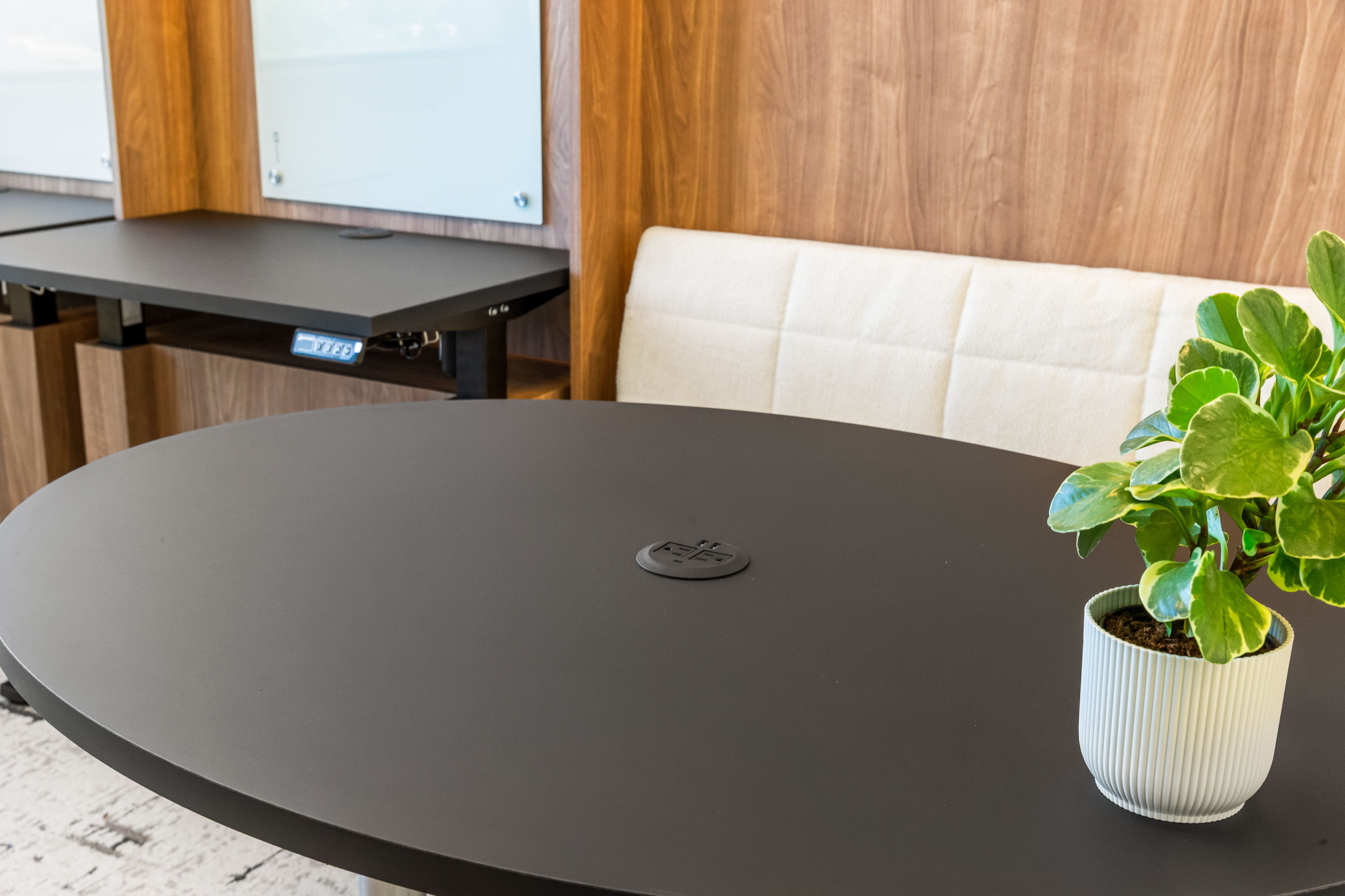 Modern office with a round black table, a small potted plant, beige cushioned bench, and a height-adjustable desk.