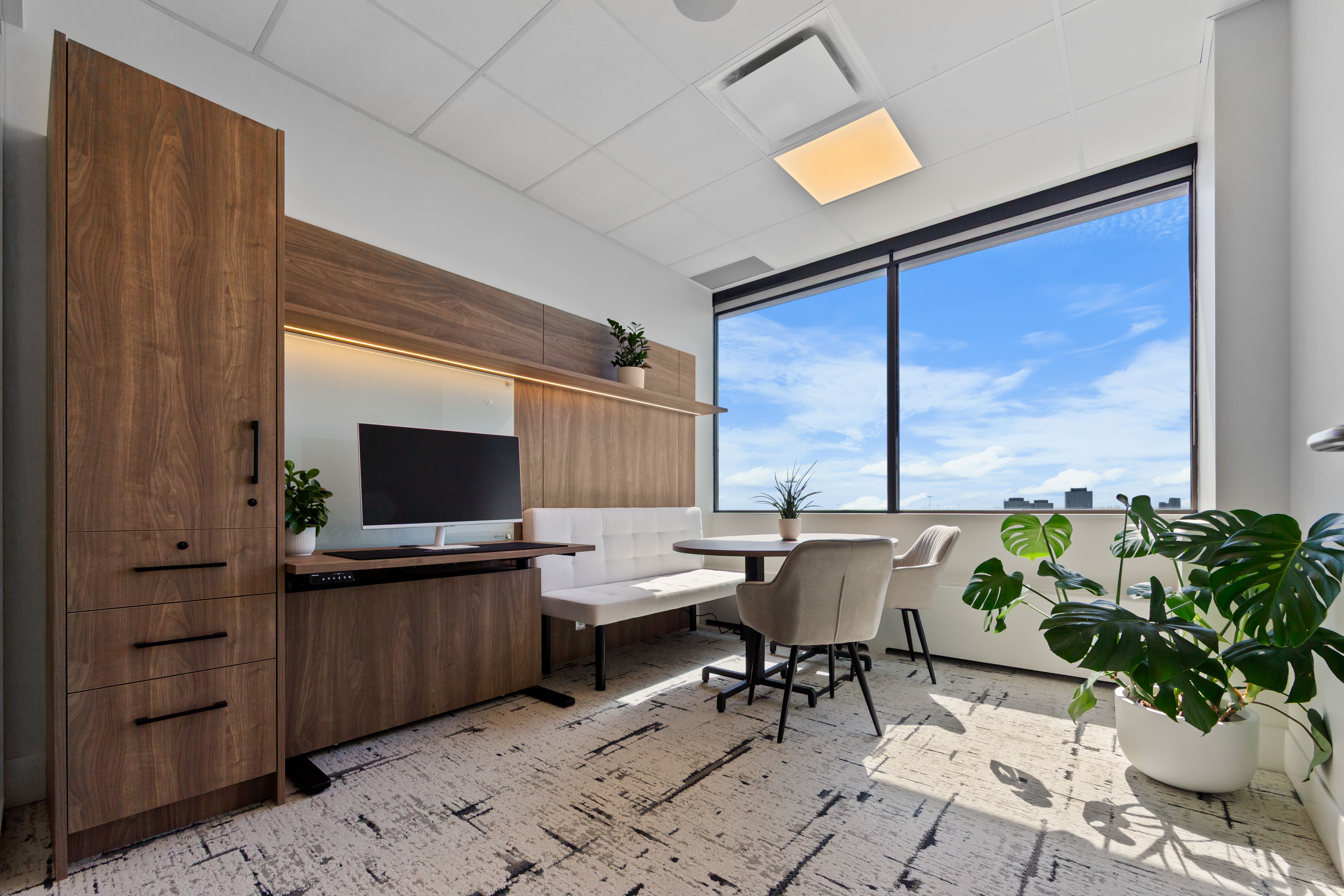 Modern office with wooden cabinetry, white bench seating, round table, two beige chairs, large window with city view, and green plants.