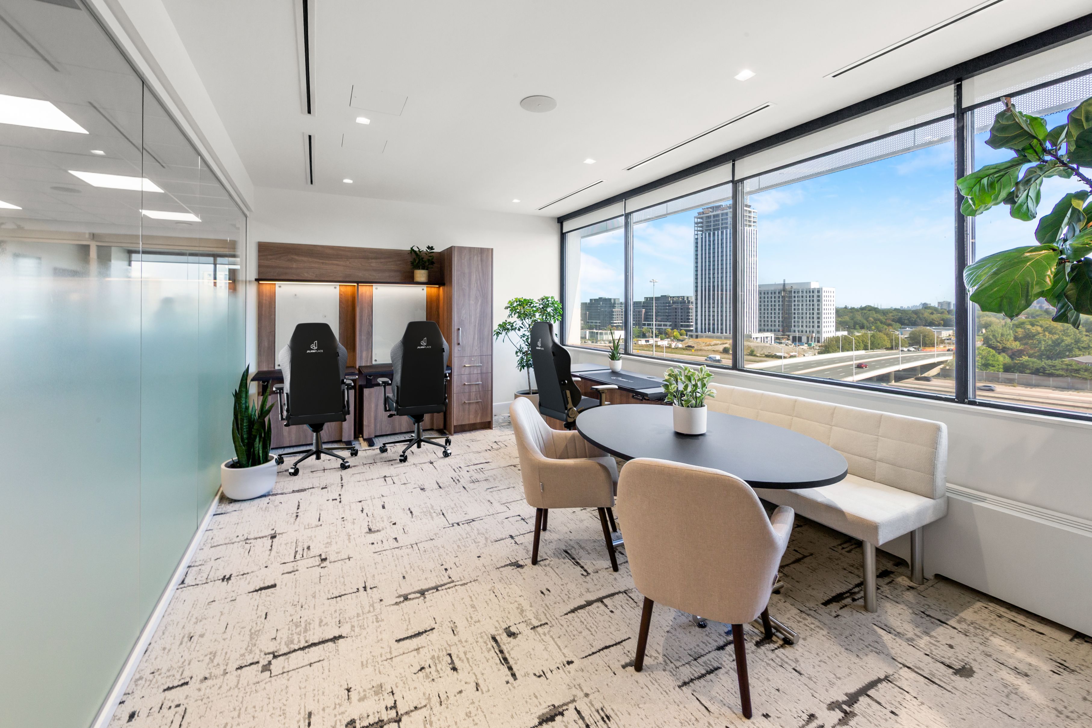 Modern office with workstations featuring black ergonomic chairs and a round table with beige chairs near large windows overlooking city buildings and a highway.