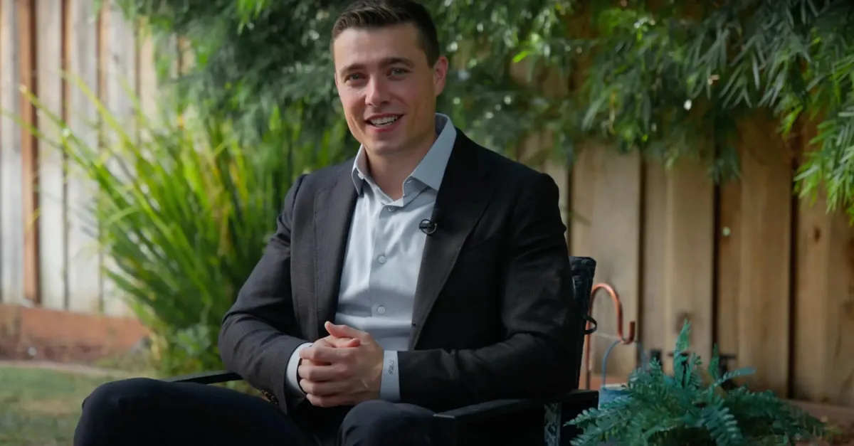 David Guardanapo, a Bay Area real estate specialist, sits outdoors in a professional black suit and light gray shirt. He is smiling warmly, seated against a backdrop of greenery and a wooden fence. The setting is relaxed and inviting, showcasing David's approachable demeanor and professionalism.