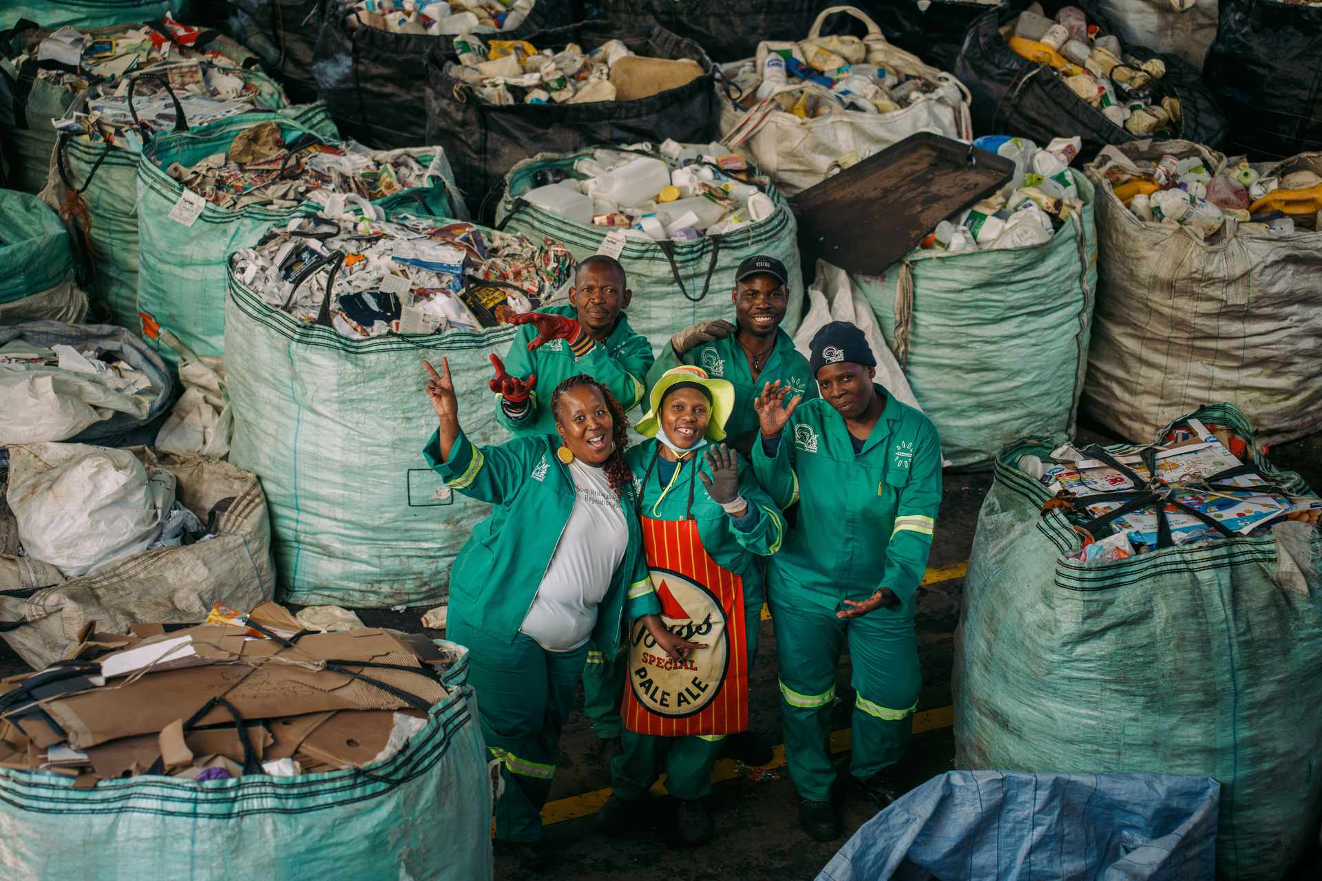 Waste pickers in green uniforms standing among bulk bags of sorted recyclable materials at a collection facility
