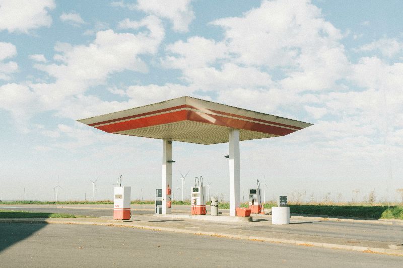 Abandoned rural gas station with four pumps under a red and white canopy, surrounded by open fields and distant wind turbines.