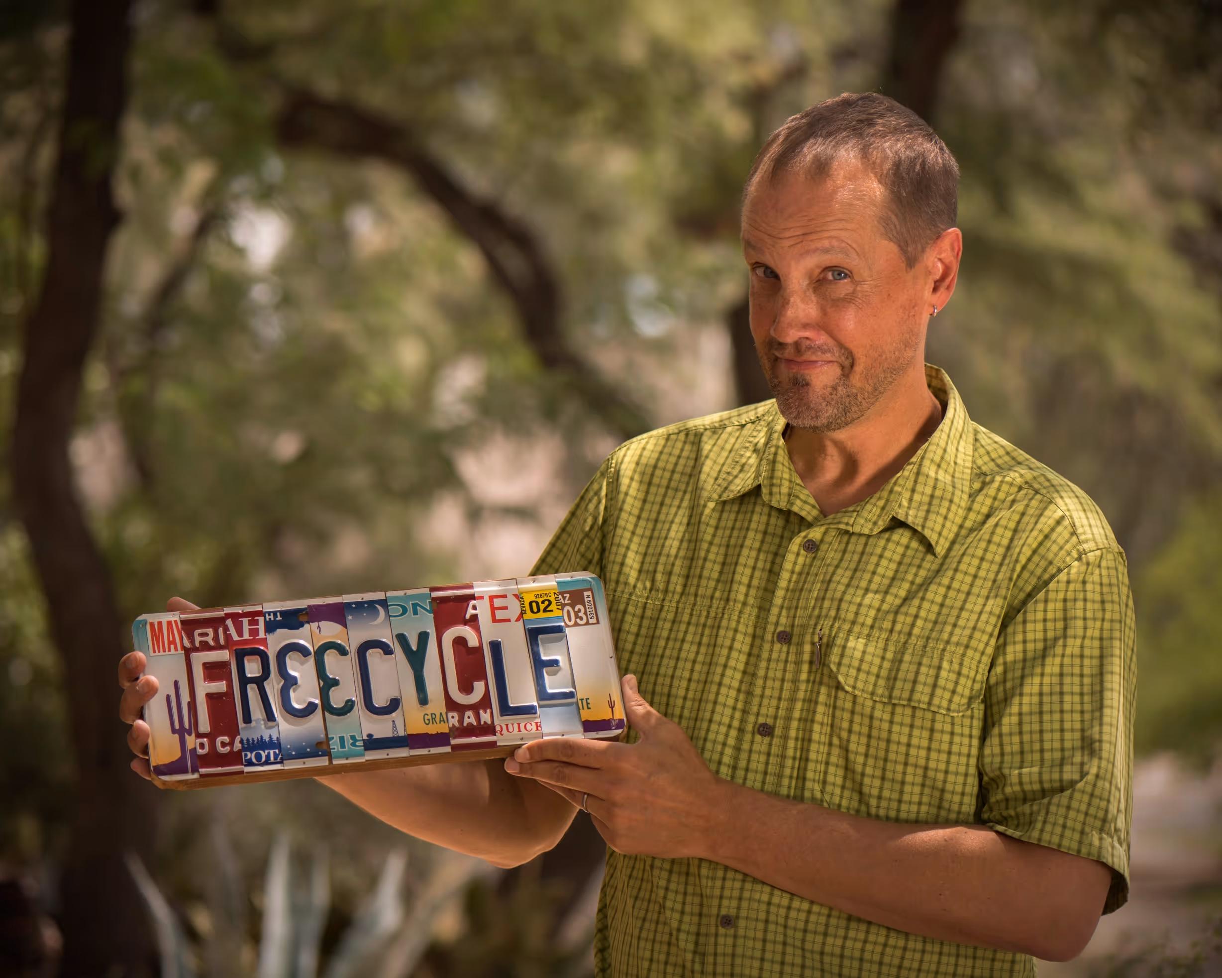 Une photo de Deron Beal, fondateur de Freecycle, tenant une plaque d'immatriculation de voiture sur laquelle est écrit FR33CYCLE. La plaque est constituée d'une combinaison de différentes plaques assemblées, chaque lettre provenant d'une plaque distincte.