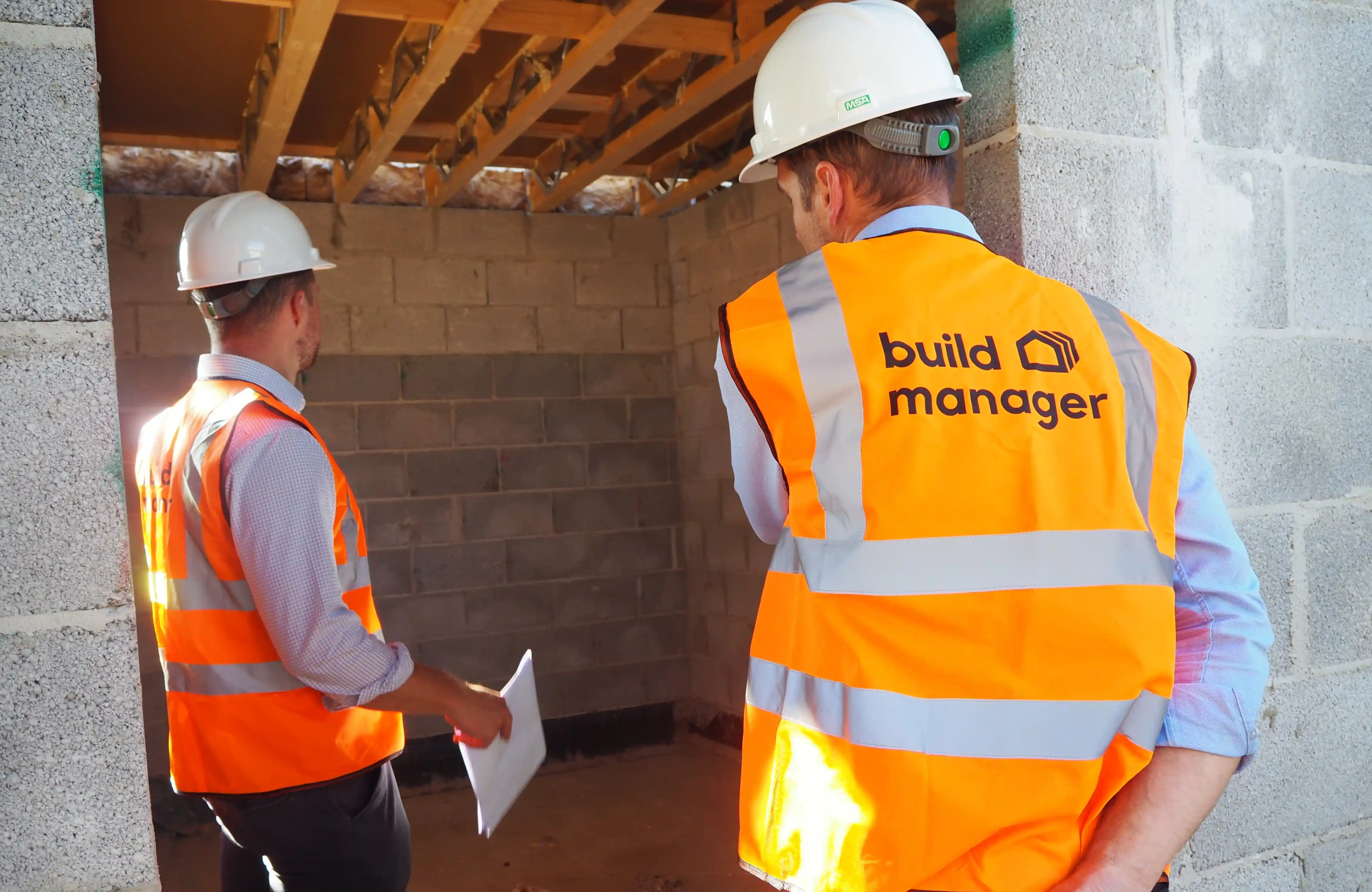 Two men in orange vests standing in front of a brick wall.