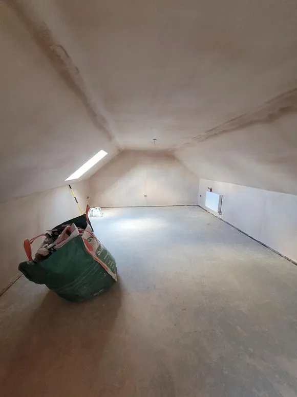 A green bag is sitting on the floor of a room that is under construction. Freshly plastered.