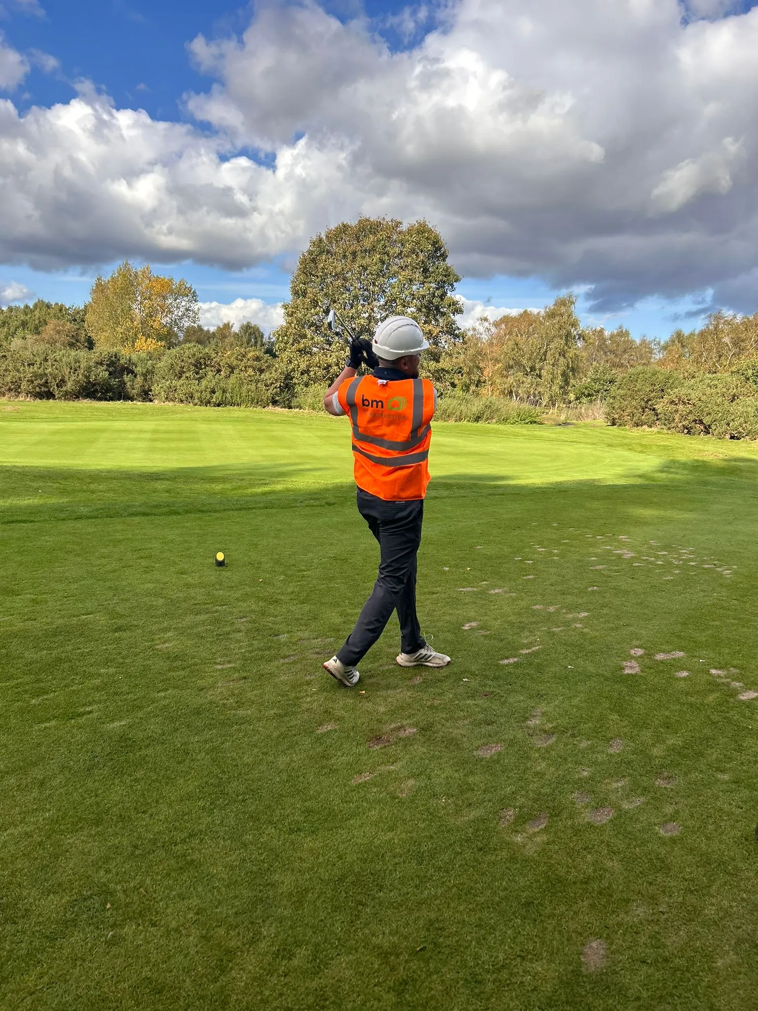 A man in an orange vest is swinging a golf club on a green course.