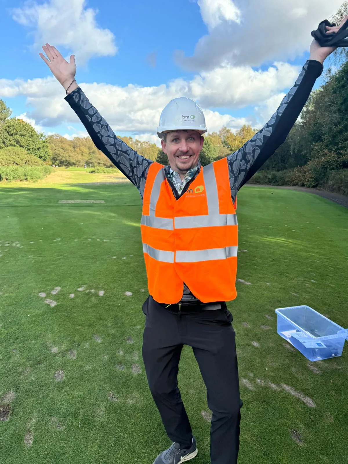 A man wearing an orange safety vest and a hard hat is standing on a golf course.