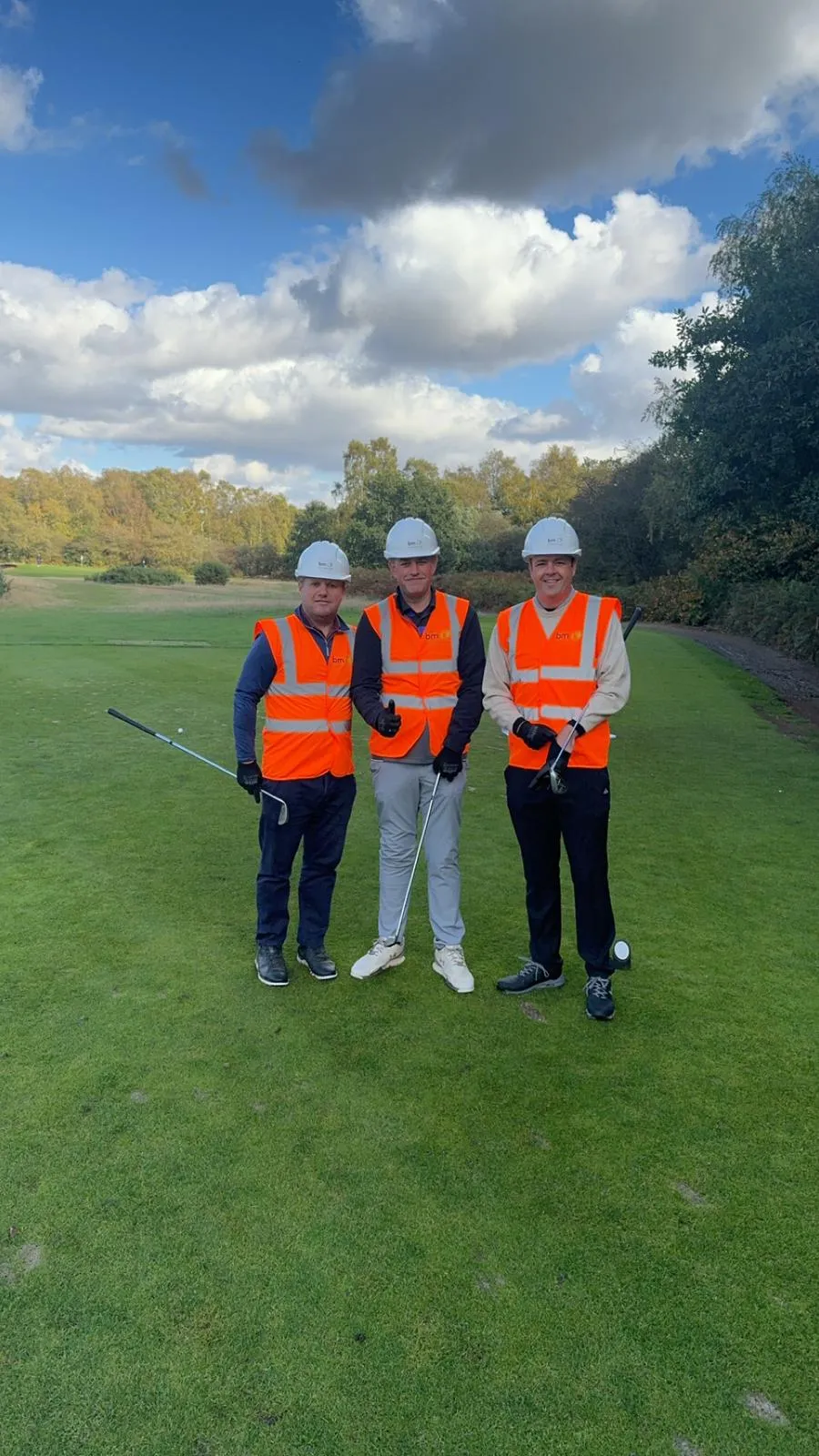 Three men wearing safety vests and holding golf clubs pose for a picture on a golf course.