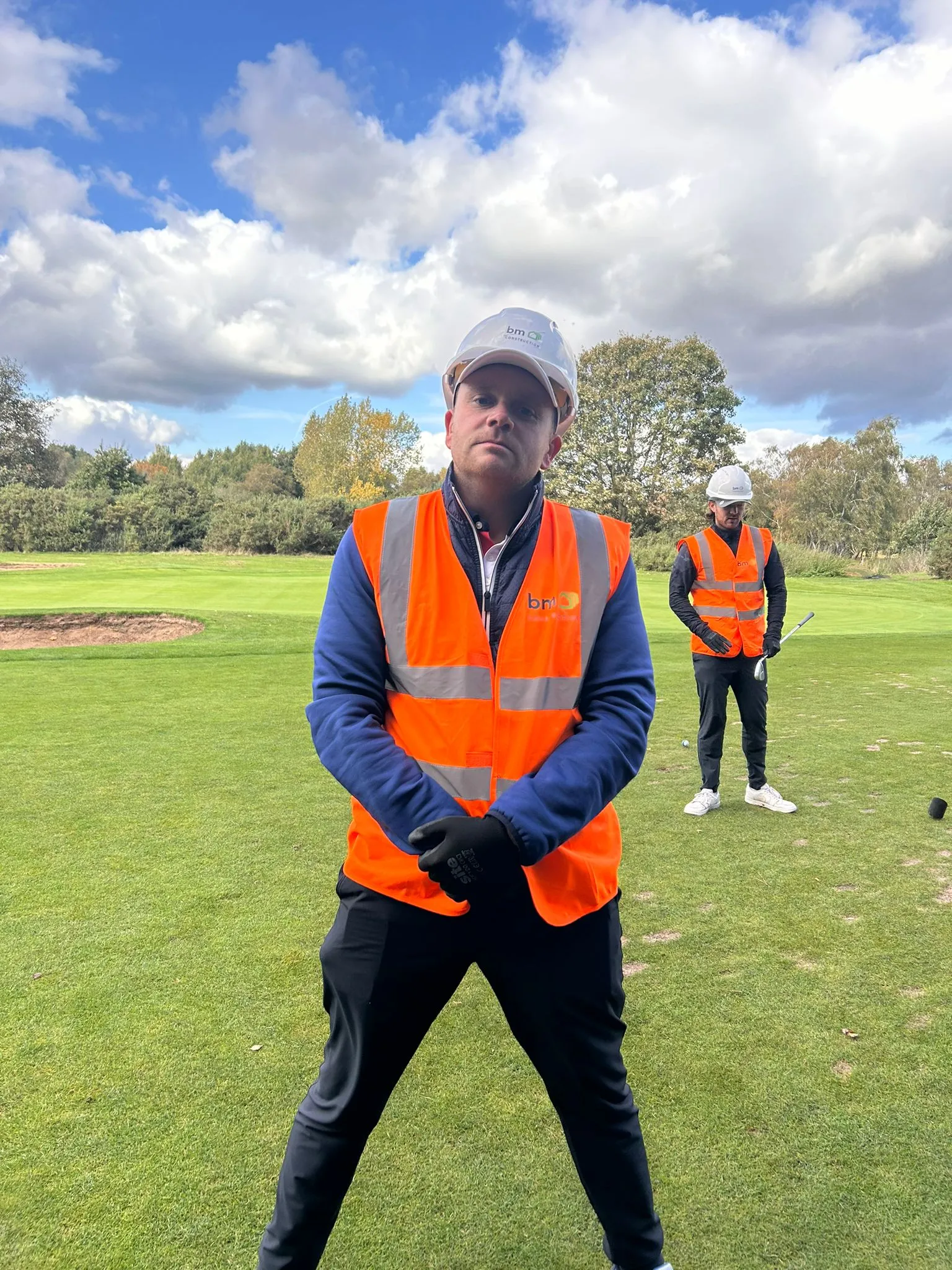 A man wearing a safety vest and a hat is standing on a golf course.