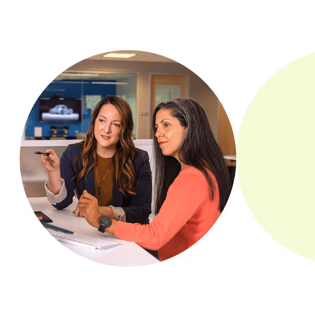 Two women in an office, one using a laptop and the other using a tablet, engaged in a collaborative discussion.

