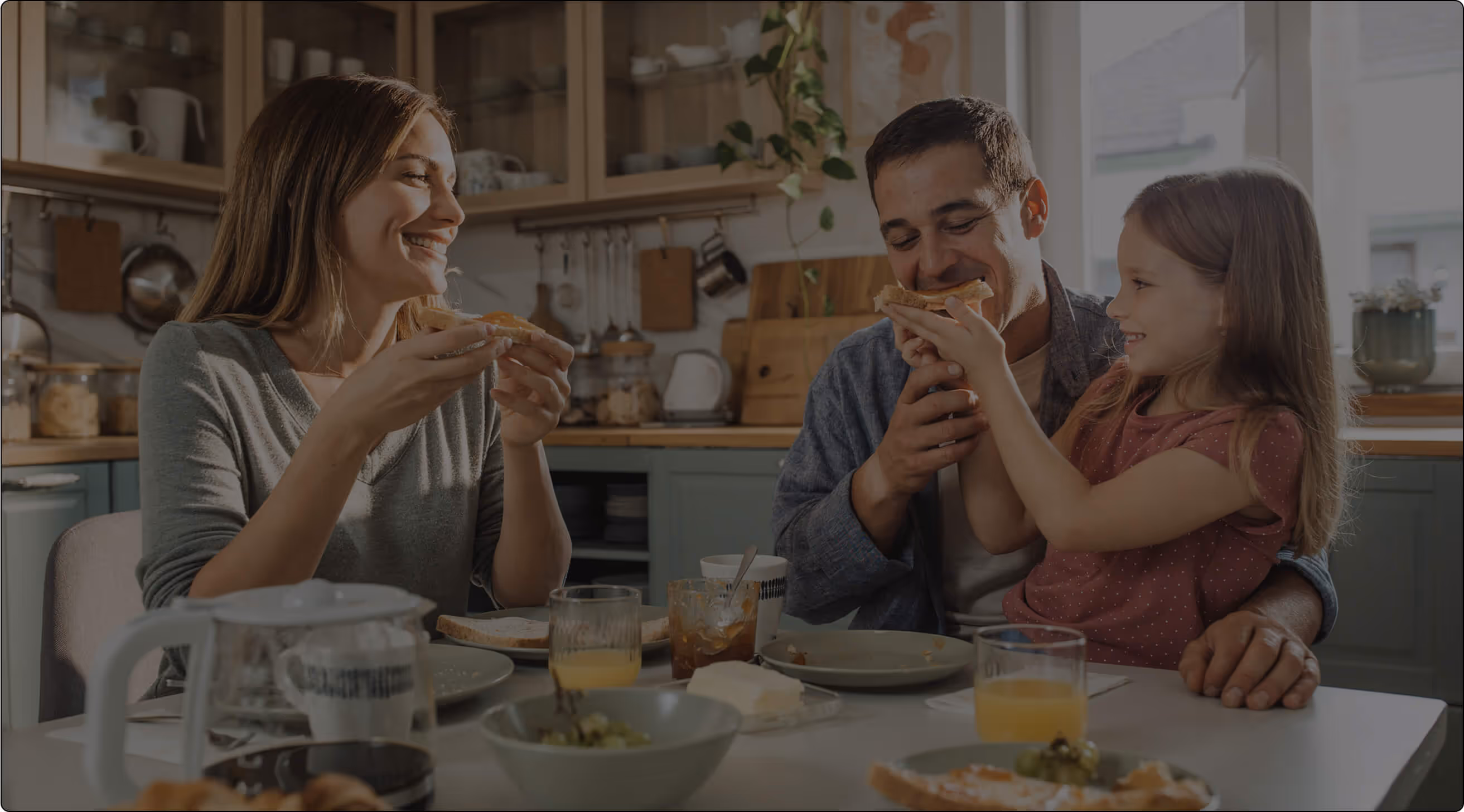 A woman, man, and young girl sit at a kitchen table eating breakfast together and smiling. The child feeds the man while the table has plates, glasses, and breakfast food.


