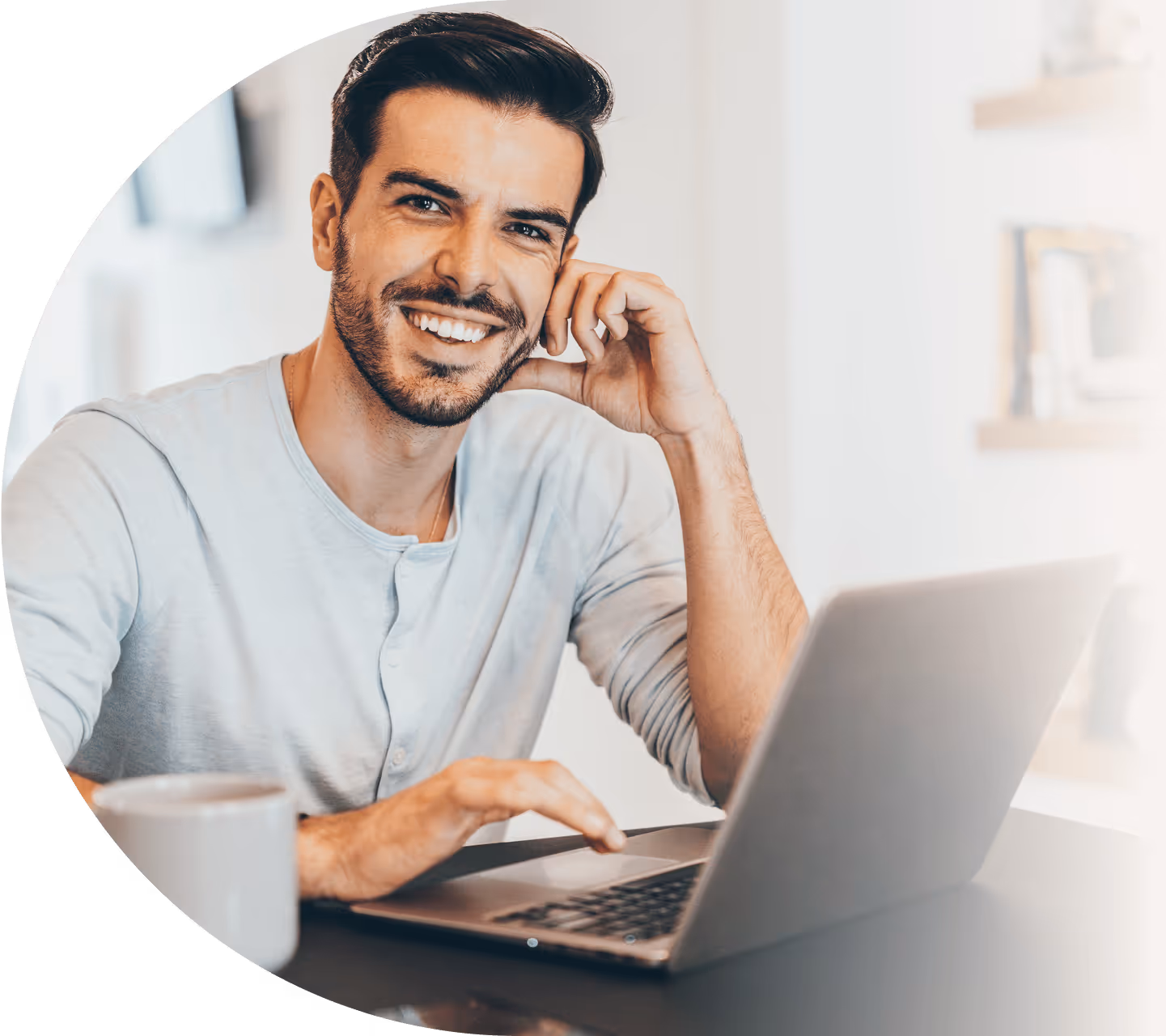 A man smiles while seated at a table, working on a laptop with a cup of coffee beside him.