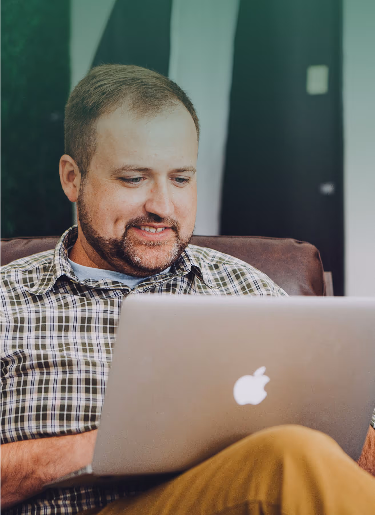 A man sitting on a couch, focused on his laptop, with a relaxed expression in a cozy living room setting.

