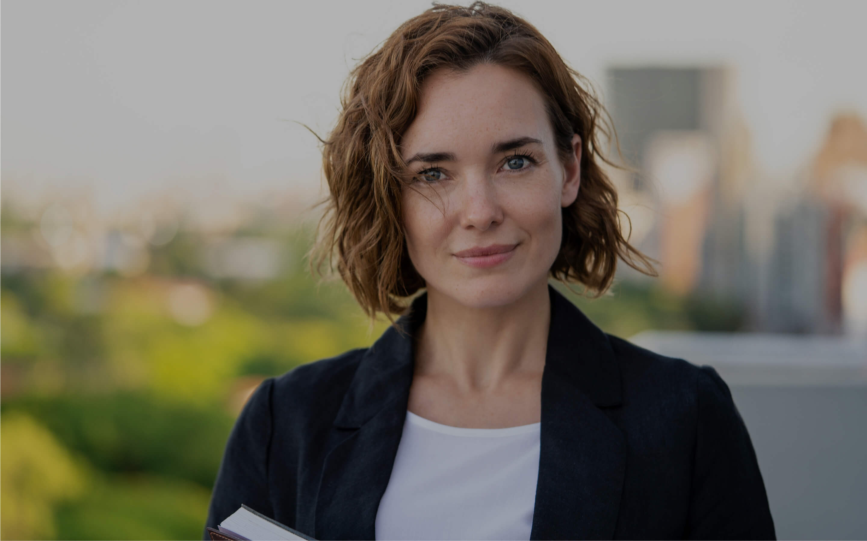 Porträt einer lächelnden Frau mit kurzen braunen Haaren im Freien, im Hintergrund verschwommene Stadtlandschaft.