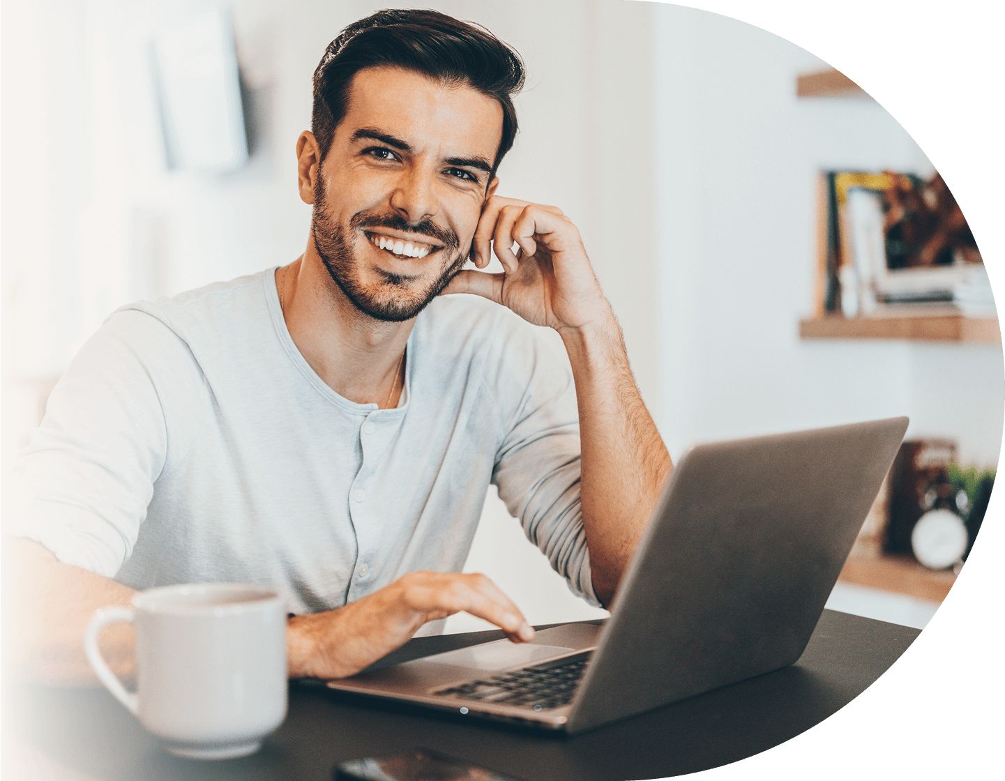 Lächelnder Mann sitzt am Tisch mit einem Laptop und einer Kaffeetasse.