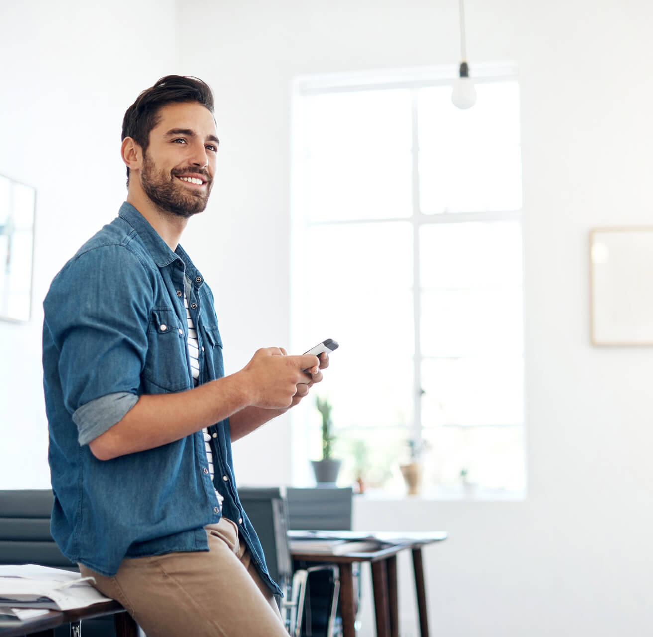 Zufriedener Mann lehnt lächelnd an einem Schreibtisch und tippt auf sein Smartphone in einem hellen Büro."