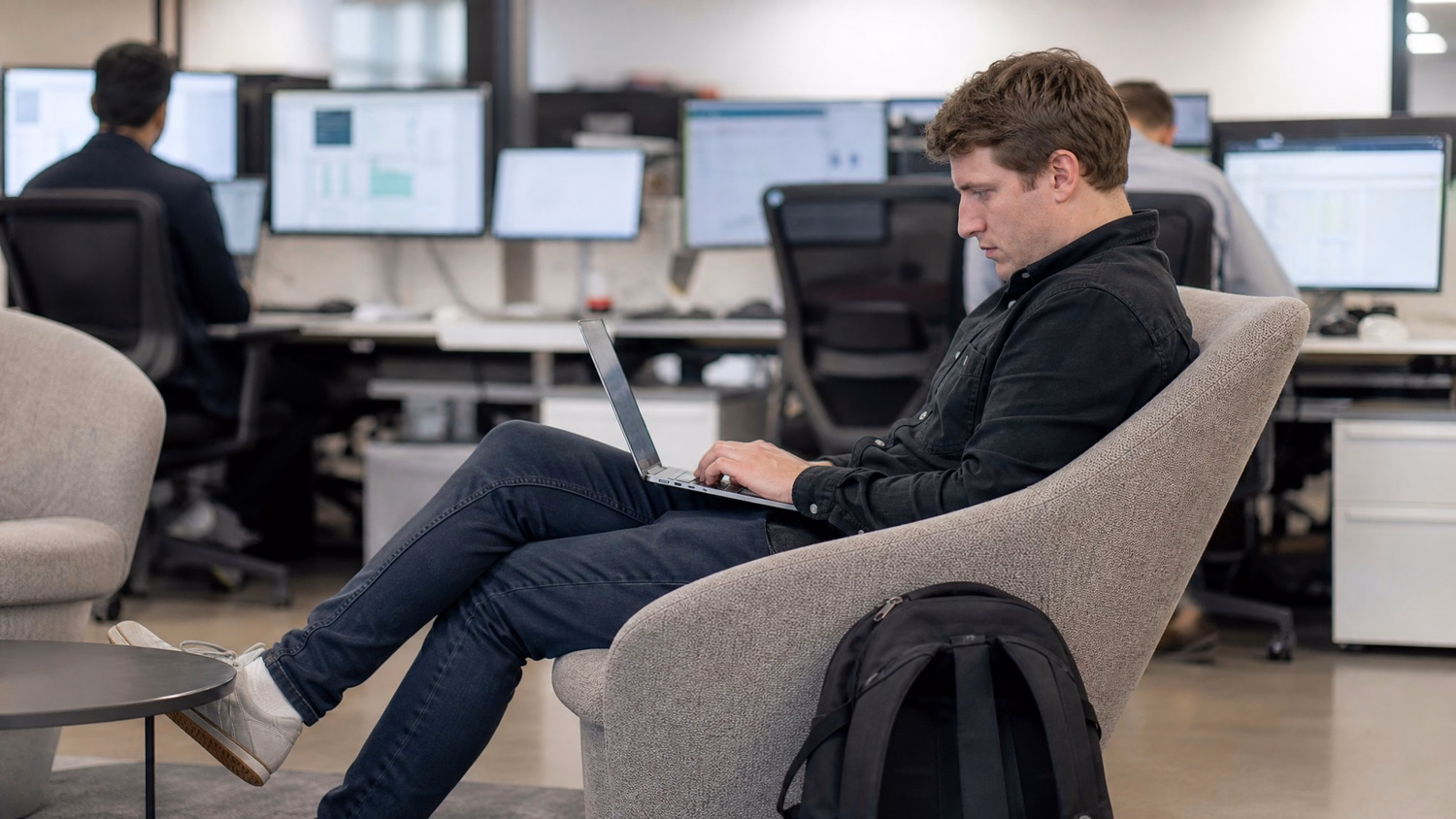 Tech professional working on laptop in lounge area, backpack nearby