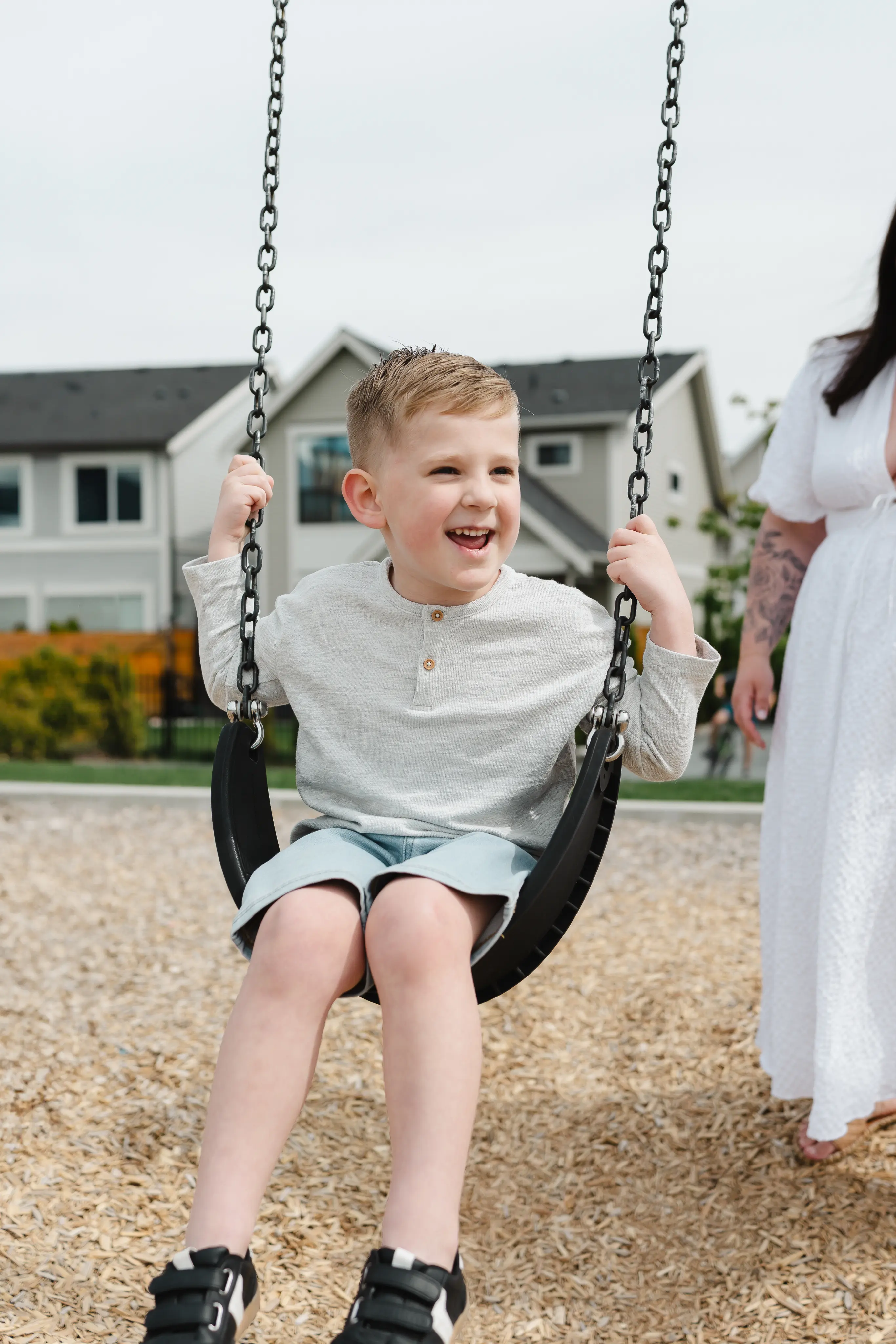 Smiling young boy in a gray shirt and shorts sitting on a swing at a playground with houses in the background.