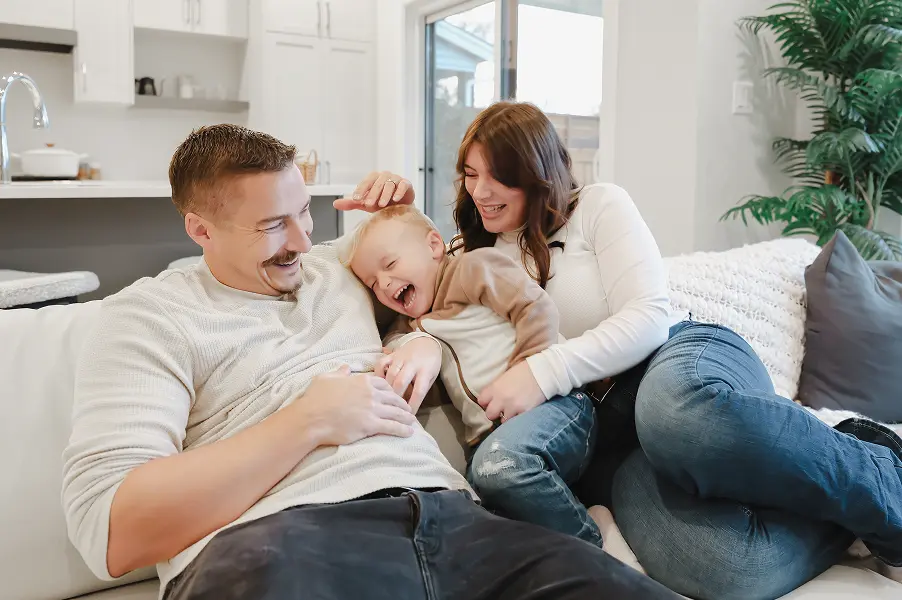 Smiling parents playfully cuddling and laughing with their young child on a living room couch.