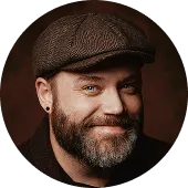 Smiling man with a beard wearing a brown flat cap and black shirt against a dark background.