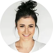 Smiling woman with dark hair in a messy bun wearing a white top against a plain light background.