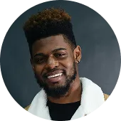 Smiling bearded man with short curly hair wearing a white jacket over a black shirt against a dark background.