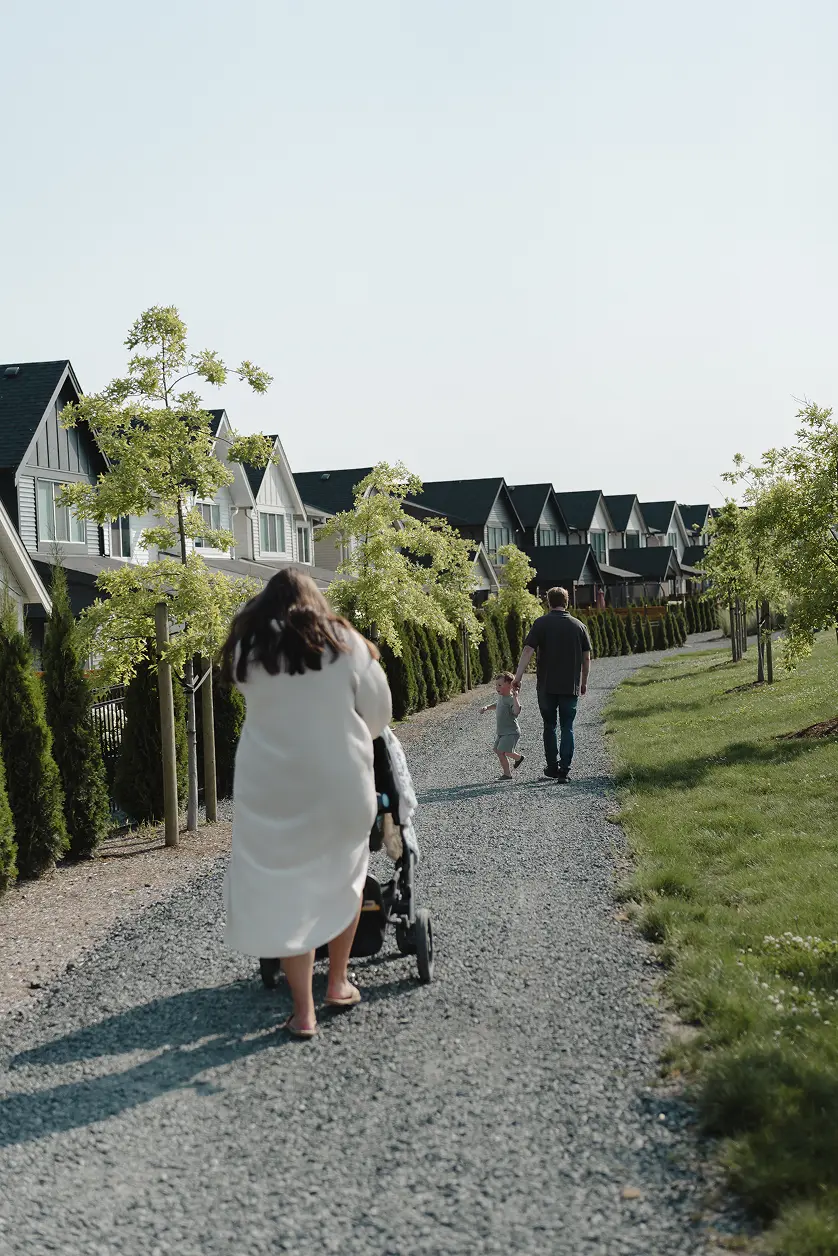Woman pushing a stroller on a gravel path lined with green trees and modern houses, with a man holding a child's hand walking ahead.