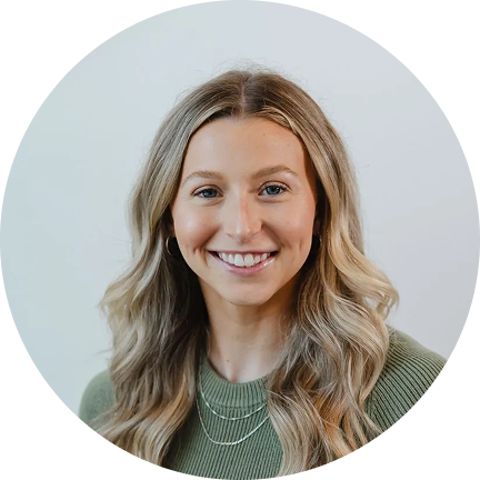 Smiling woman with shoulder-length blonde hair wearing a green top in front of a plain light background.