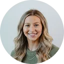 Smiling woman with shoulder-length blonde hair wearing a green top in front of a plain light background.