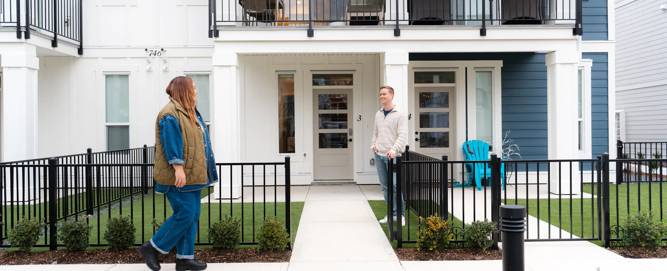 Two people standing outside separate entrances of a modern white and blue townhouse with black metal fences and small bushes.