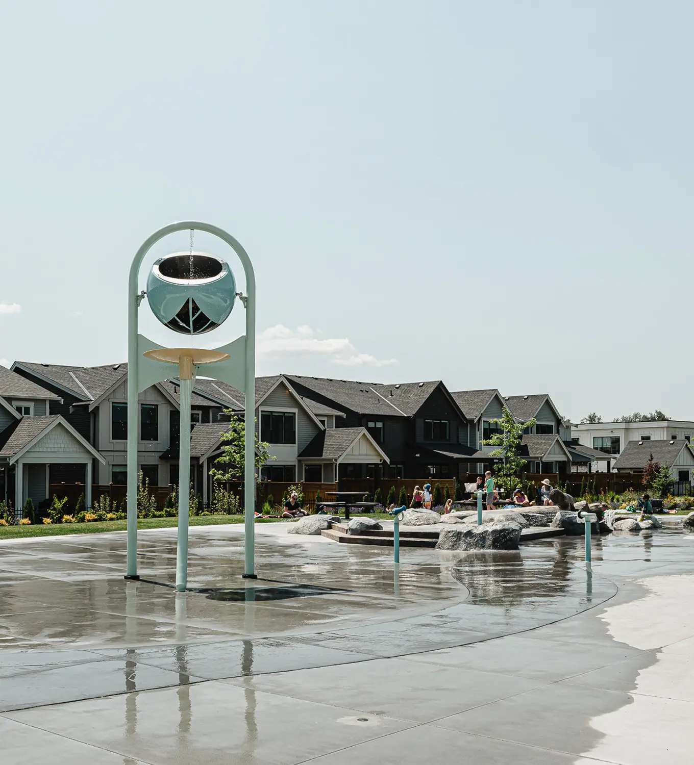 Children playing in a wet urban splash pad with water features and houses in the background.