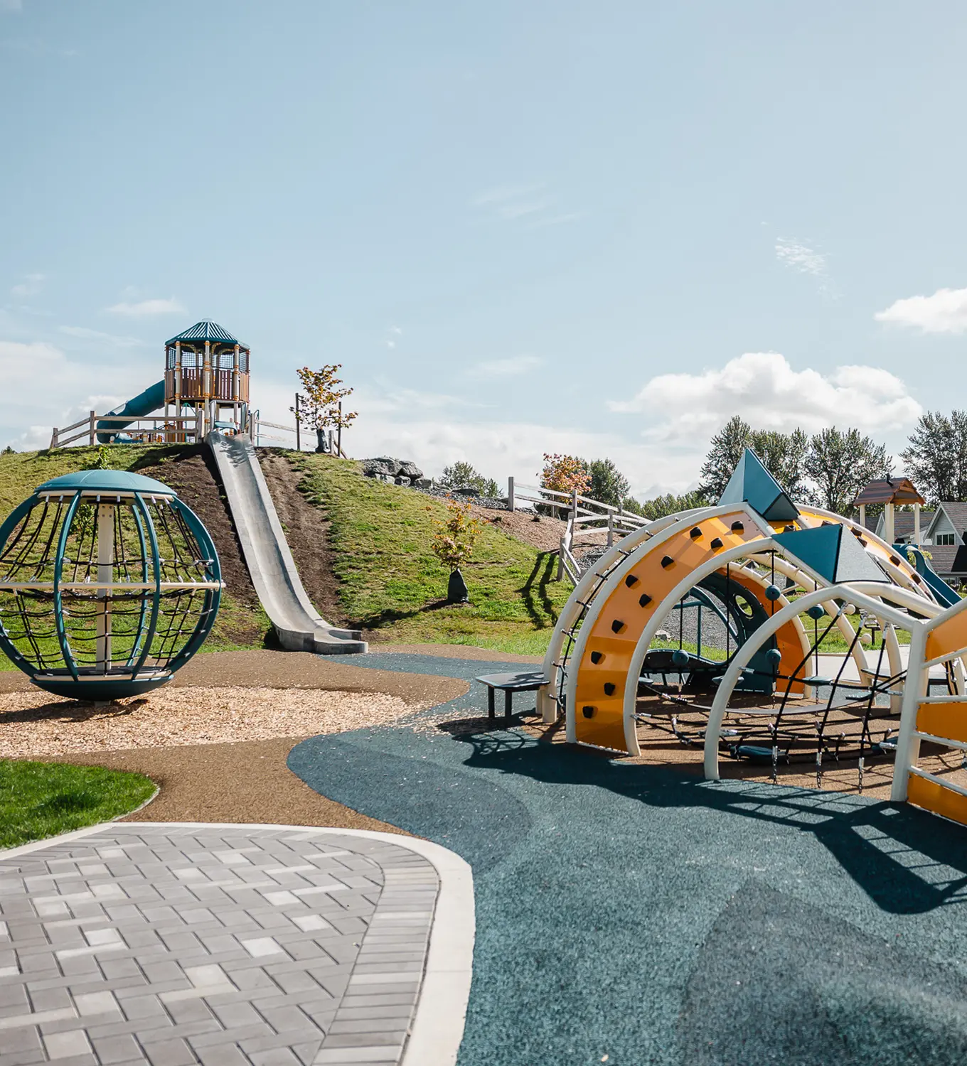 Playground with a tall slide on a grassy hill, a spherical climbing structure, and yellow and blue climbing arches under a clear sky.