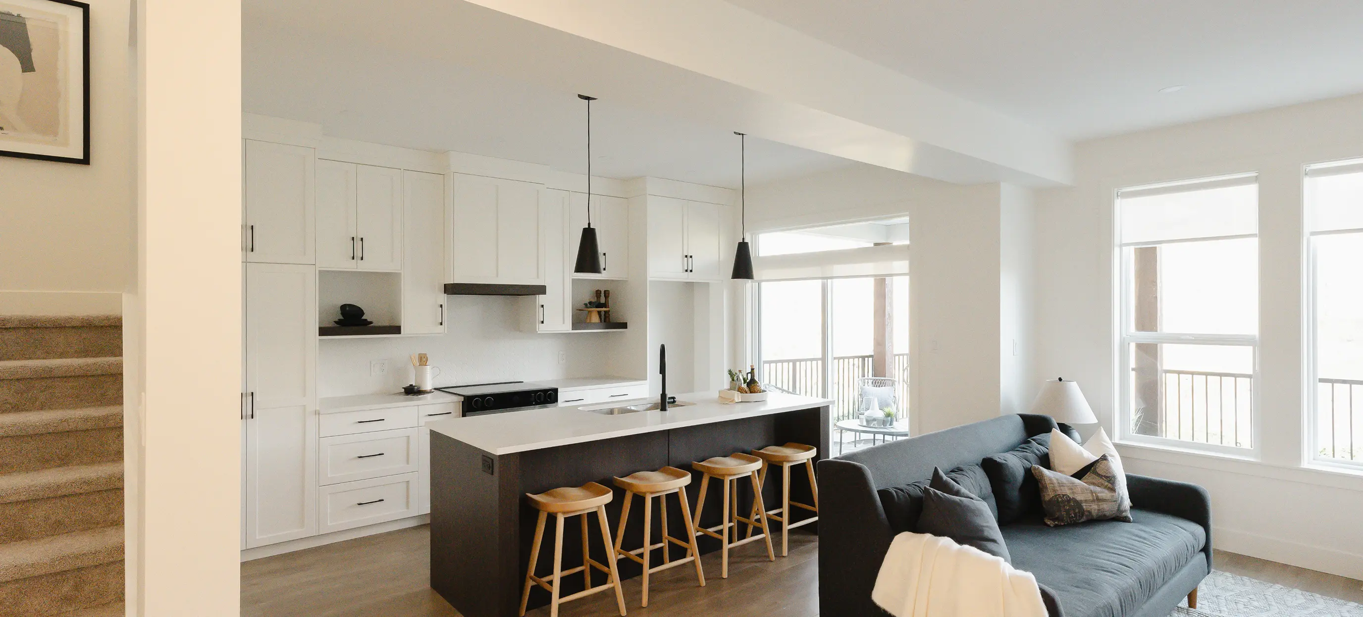 Modern open kitchen with white cabinets, a dark island with four wooden stools, and a living area featuring a dark gray sofa near large windows.