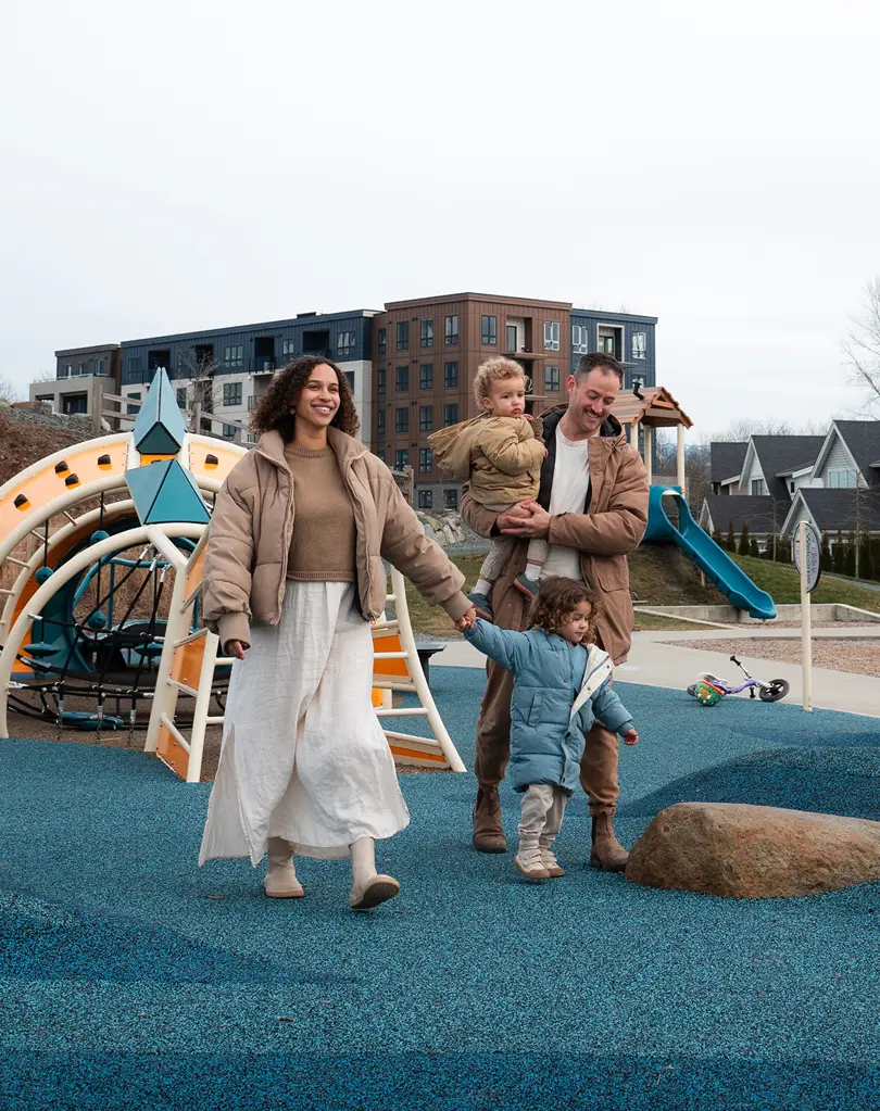 Family of four at a playground with blue rubber flooring and modern apartment buildings in the background.