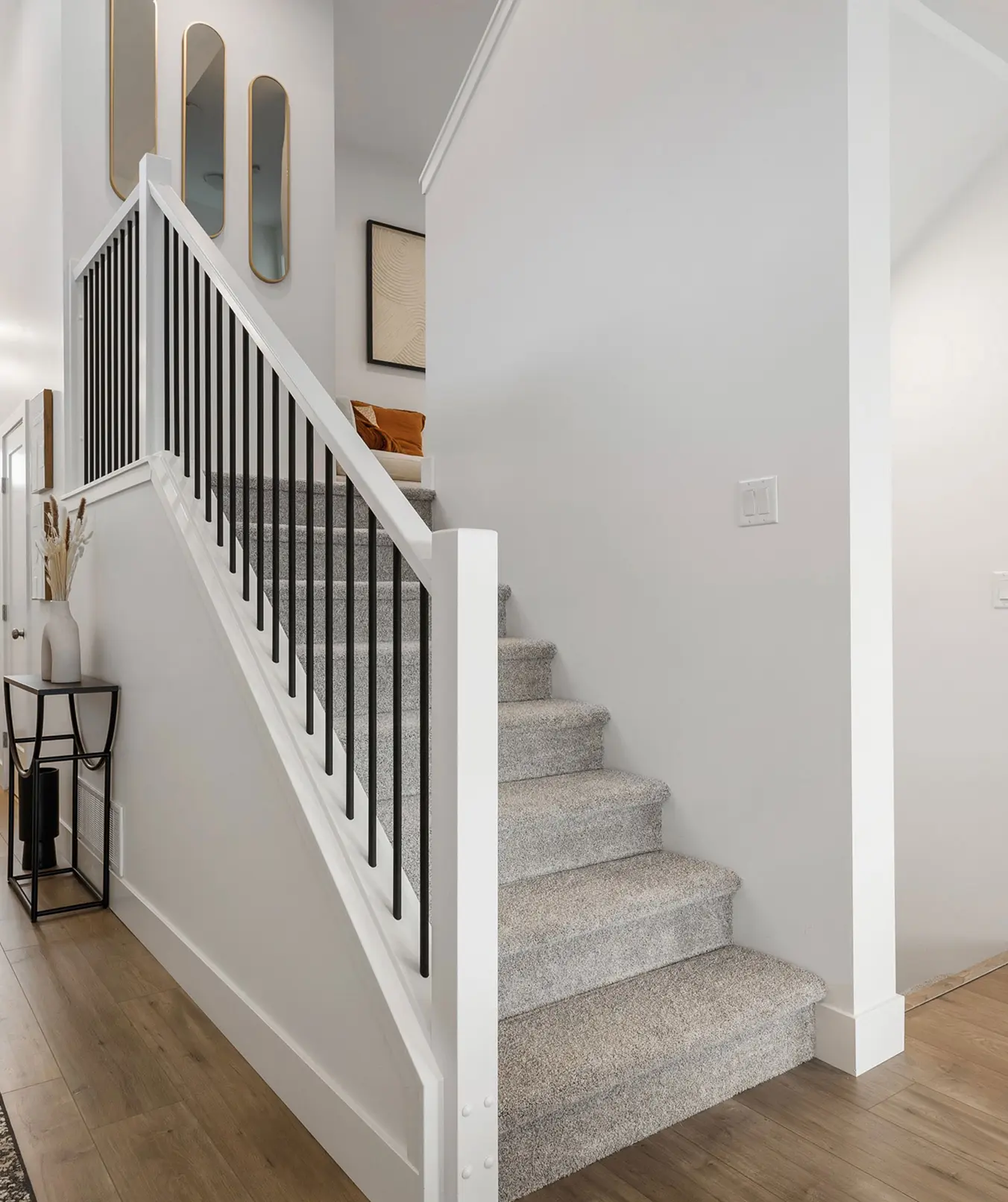 Indoor staircase with grey carpet, white railing with black balusters, wooden floor, and decorative mirrors on the wall.