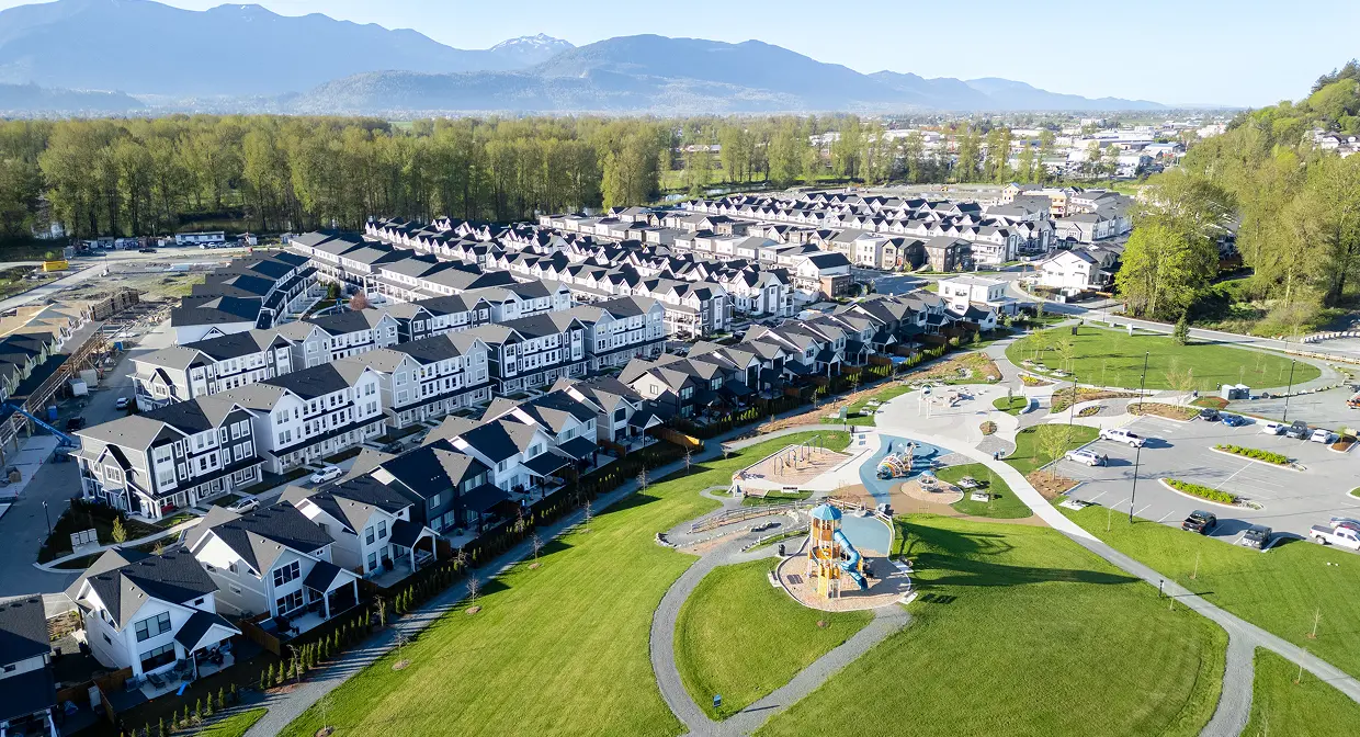 Aerial view of a suburban housing development with rows of modern townhouses, a green park with a playground, and a parking area, set against a backdrop of forest and mountains.