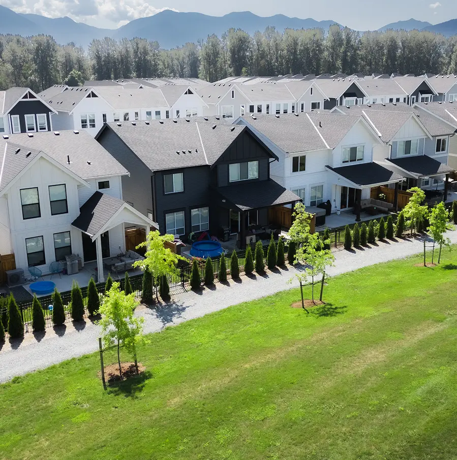 A row of modern two-story suburban houses with light and dark siding, small fenced backyards, and a green grassy area with young trees in front.