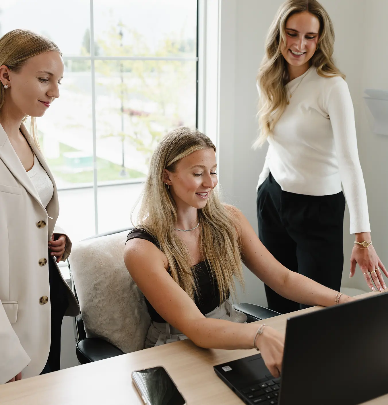 Three women collaborating at a desk with a laptop and smartphone, smiling and engaged.