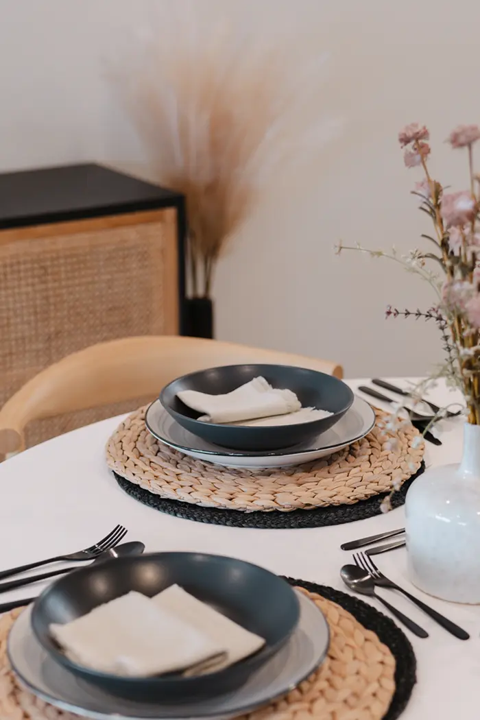 Modern dining table set with black bowls, white plates, beige napkins, woven placemats, and floral centerpiece in a white vase.