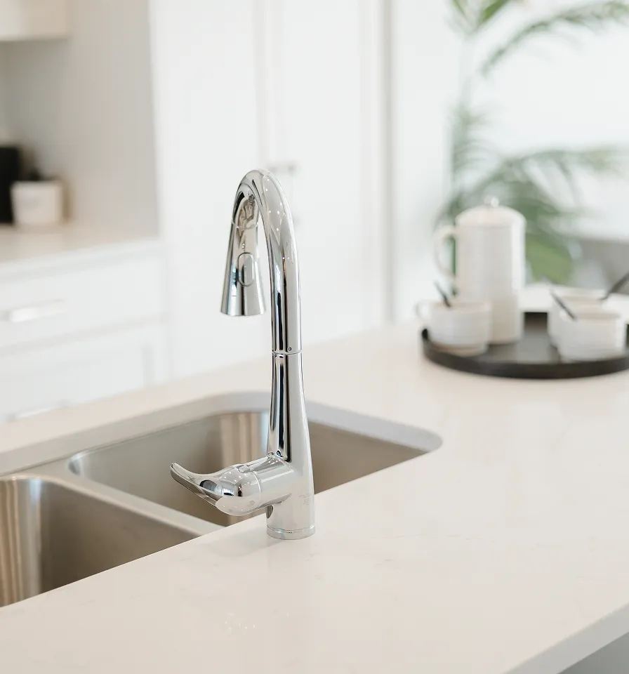 Modern chrome kitchen faucet installed on a white marble countertop with a double stainless steel sink and a blurred tray with dishes in the background.