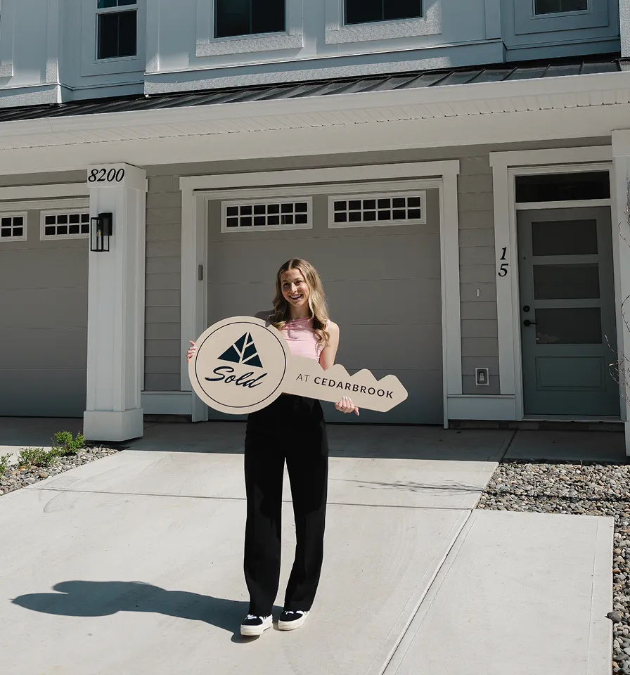 Smiling woman holding a large wooden key sign reading 'Sold at Cedarbrook,' standing in front of a modern house with gray garage doors.