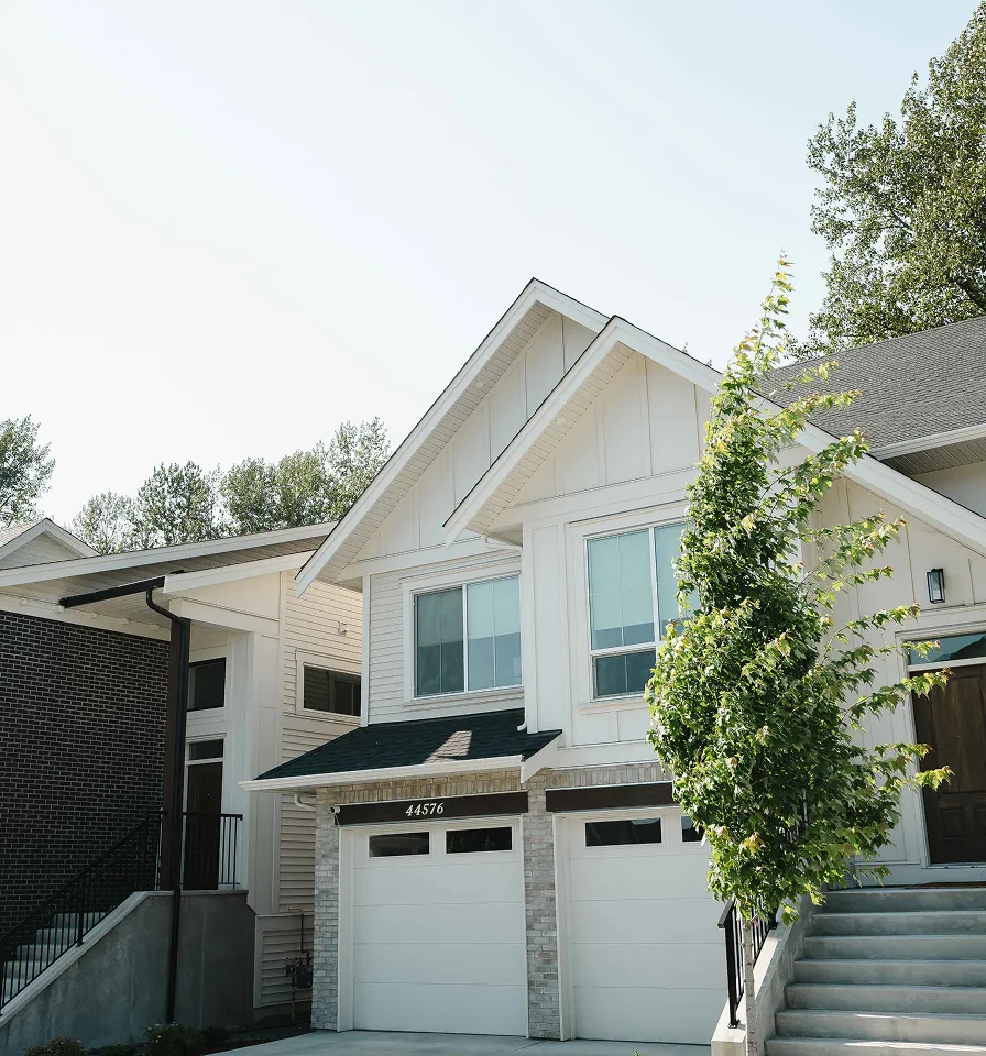 White suburban house with two garage doors, a tree near the stairs, and surrounding greenery on a clear day.