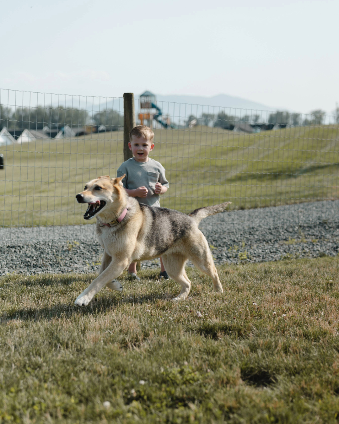 Young boy swinging on a playground swing with a woman in a white dress standing nearby.